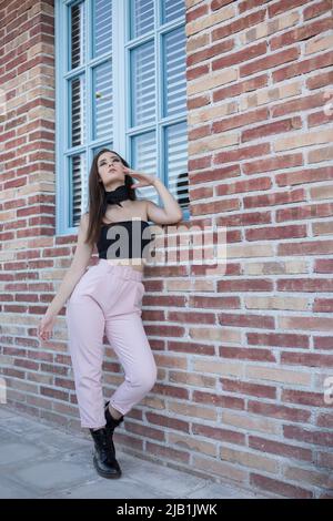 Vista laterale del corpo della giovane donna in un abito alla moda in piedi vicino alla parete di mattoni e toccare i capelli mentre si guarda la macchina fotografica Foto Stock