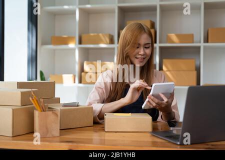 Giovane donna piccola impresa proprietario online shopping a casa. Confermare gli ordini da parte dei clienti con i telefoni cellulari preparare il prodotto pacchetto in background Foto Stock