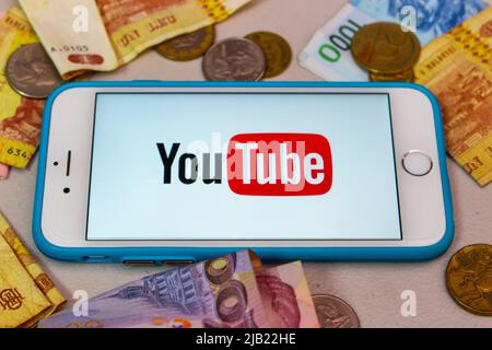 Il logo di YouTube su un iPhone con denaro casuale (banconote e monete). YouTuber, Vtuber e influencer business per il concetto di monetizzazione Foto Stock