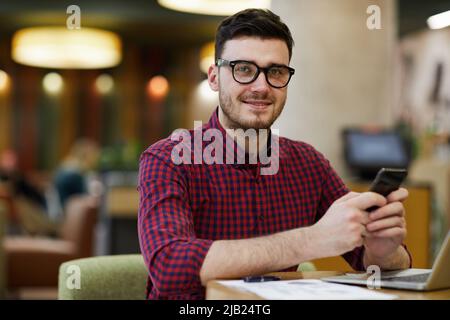 Sorridente, giovane e intelligente manager maschile in maglia a scacchi seduta al tavolo e utilizzando lo smartphone per le comunicazioni online mentre si lavora in un bar Foto Stock