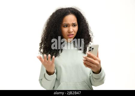 Deluso giovane afroamericana che tiene smartphone e guardando lo schermo del telefono, signora ha ricevuto cattiva notifica o messaggi, si sente sconvolto, shrugging, isolato Foto Stock