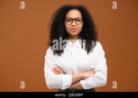 Felice giovane donna multirazziale con le braccia ripiegate sta vicino spazio vuoto. Ragazza allegra donna indossando abiti casual con le braccia incrociate isolato color marrone pastello sfondo Foto Stock