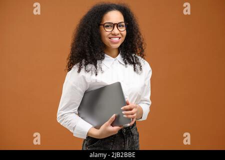 Donna che trasporta un computer portatile, sorridente. Giovane afroamericana con acconciatura afrofroche indossa abbigliamento casual elegante e occhiali eleganti che si erge isolato sul marrone. Dipendente d'ufficio, concetto di studente Foto Stock