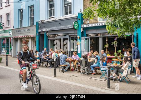 Un uomo in bicicletta passa davanti a persone seduti fuori mangiare e bere all'Exmouth Market, Clerkenwell, Londra EC1 Foto Stock