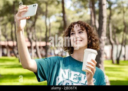 Allegra donna rossa con tee verde e in piedi sul parco cittadino, all'aperto con selfie e tazza da caffè da asporto. Autoritratto per sociale m Foto Stock