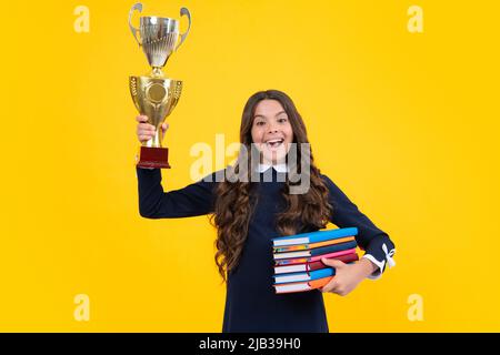 Studentessa entusiasta in divisa scolastica che celebra la vittoria con il trofeo. Premio vincente per l'adolescente su sfondo giallo. Libretto di custodia per bambini con Foto Stock