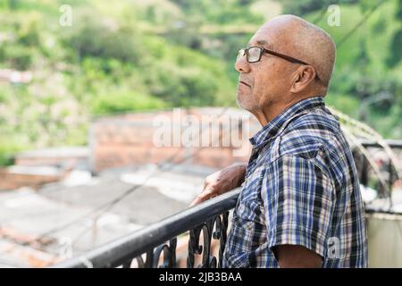 primo piano dell'anziano latino con pelle marrone che si alza sul balcone della sua casa guardando verso il cielo, pensando alla sua vita Foto Stock