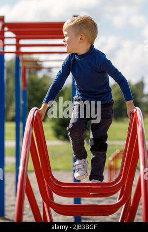 Un ragazzino si arrampica e si appende su una scala di metallo a forma di arco nel parco giochi. Foto Stock