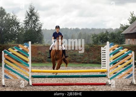 Sport equestre. La giovane ragazza corre a cavallo in campionato, salta attraverso la barriera con cavallo marrone ciliegia. Foto Stock