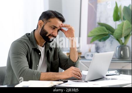 L'uomo arrabbiato arabo o indiano, stanco, sovraccaricato di lavoro in un computer portatile, guarda tristemente allo schermo, è sottolineato a causa di un lavoro non fatto in tempo, un progetto fallito, una startup, ha bisogno di una vacanza Foto Stock