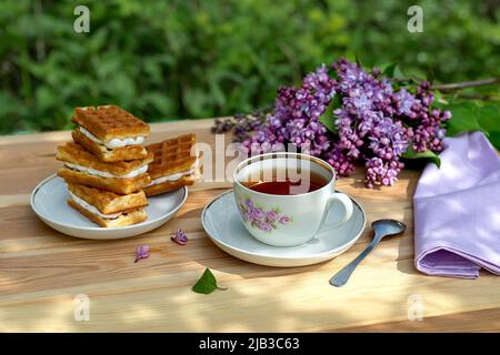 Festa del tè con cialde viennesi e un ramo di lilla su un tavolo di legno con ombre sullo sfondo del giardino in una giornata estiva soleggiata. Foto Stock