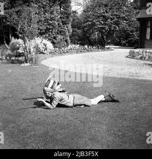 1950s, storico, fuori in giardino, un ragazzo che gioca, fingendo di essere un indiano nativo o rosso, adagiato sull'erba, con il suo fucile giocattolo, cappello di piuma e volto dipinto, Inghilterra, Regno Unito. Nella Gran Bretagna del dopoguerra, giocare a cowboy e indiani era un popolare gioco di make-credi per i bambini piccoli. Foto Stock