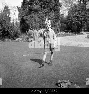 1950s, storico, fuori in giardino, un ragazzo in piedi sull'erba, giocando, fingendo di essere nativo o un indiano rosso, con una giacca in costume, un arco sulla spalla, un cappello di piuma e volto dipinto, Inghilterra, Regno Unito. Nella Gran Bretagna del dopoguerra, giocare a cowboy e indiani era un popolare gioco di make-credi per i bambini piccoli. Foto Stock