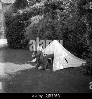 1950s, storico, in giardino, seduto fuori dalla sua tenda, un ragazzo che gioca, fingendo di essere un nativo o un indiano rosso, con un cappello di piuma e volto dipinto, Inghilterra, Regno Unito. Un arco fatto in casa è sull'erba accanto alla tenda. Nella Gran Bretagna del dopoguerra, giocare a cowboy e indiani era un popolare gioco di make-believe per i bambini piccoli e vestirsi in temi occidentali era una cosa divertente da fare. Foto Stock