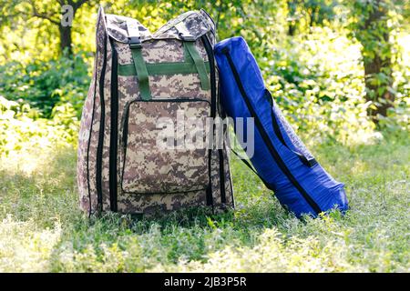 Zaino militare defocus e tenda blu o sacco a pelo. Borsa esercito su sfondo verde erba vicino albero. Zaino militare mimetizzazione. Turismo summ Foto Stock