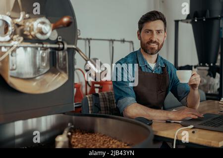 Barista sorridente che beve caffè seduto accanto alla macchina per tostare il caffè Foto Stock