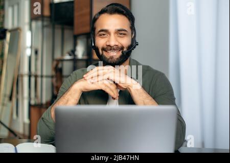 Ritratto di positivo operatore di call center arabo o indiano, consulente hotline, mentore, con cuffie, conduce la consultazione online con i clienti o lezioni online con lo studente, guarda la fotocamera, sorrisi Foto Stock