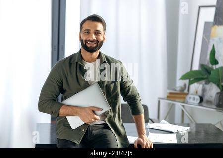 Ritratto di un uomo indiano o arabo di successo carismatico positivo, amministratore delegato della società, freelance o designer creativo che tiene il laptop, in piedi nel suo ufficio moderno, guardando la macchina fotografica, sorridente amichevole Foto Stock