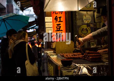 Jioufen, Taiwan - 19 2019 dicembre: Via dello shopping di Jioufen sotto la pioggia. Giovane coppia curiosamente guardando il negozio di salsiccia da asporto. Torta di fagioli rossi (torta di ruote) Foto Stock