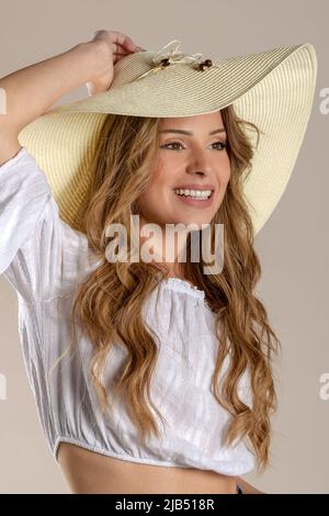 Giovane donna felice in blusa bianca con capelli lunghi e leali che toccano il grande cappello estivo e che guarda via con il sorriso su sfondo beige Foto Stock