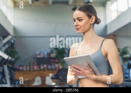 Ritratto interno di una giovane donna millenaria bianca con capelli leccati e reggiseno sportivo grigio che tiene un tablet, creando un programma di allenamento per i suoi clienti. Foto di alta qualità Foto Stock