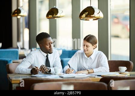 Contenuti pensivi giovani colleghi multietnici in camicie bianche seduti a tavola in un bar e lo scambio di strategie di business mentre si guarda la carta Foto Stock