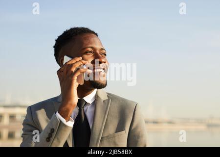Sorridente giovane uomo d'affari afro-americano con barba che parla sul cellulare mentre si cammina in città Foto Stock
