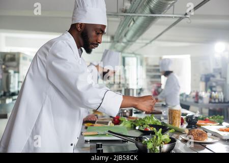 Esperto dell'industria alimentare che aggiunge erbe fresche verdi per migliorare il gusto del piatto. Esperto di gastronomia che indossa uniforme di cucina mentre prepara deliziosi piatti gourmet per il servizio cena al ristorante. Foto Stock