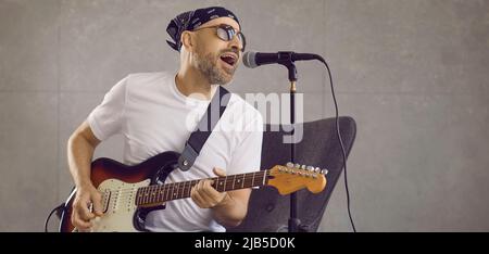 Musicista rock in bandana e occhiali da sole che suonano chitarra elettrica e cantano in studio Foto Stock