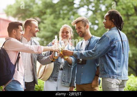 Gruppo di giovani allegri interrazziali in piedi nel parco della città e bottiglie di clinking mentre beve all'amicizia Foto Stock