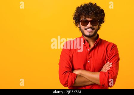 Ritratto di felice soddisfatto bel giovane uomo in camicia in piedi con le braccia incrociate e guardando la macchina fotografica con il sorriso toothy. Studio interno girato su sfondo arancione Foto Stock