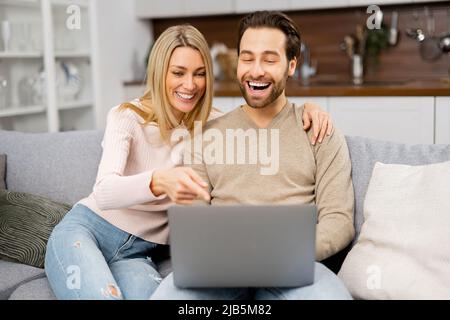 Coppia serena che passa il tempo insieme a casa, usando il laptop per qualcosa. Giovane moglie che punta sullo schermo mentre si riposa sul divano in appartamento moderno con suo marito, guardando il film insieme Foto Stock