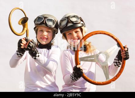 Le sorelle gemelle immaginano che stiano guidando un'auto da corsa. Indossano caschi d'epoca, occhiali e usano un vecchio volante timer e corno per i puntelli. Foto Stock