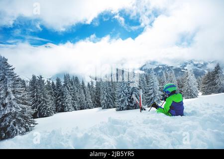 Il bambino si siede nella neve con lo sci sopra la foresta di abeti dopo la nevicata pesante Foto Stock