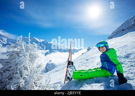 Ragazzo con sci alpino seduto nella neve sopra le cime di montagna Foto Stock