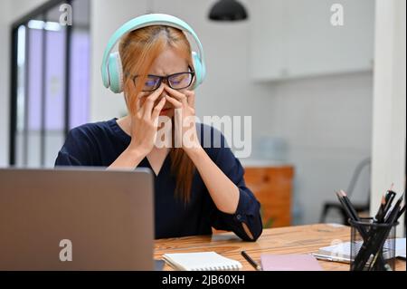 Triste donna teen che indossa cuffie wireless, ascoltando la musica dallo smartphone mentre si siede nel suo spazio di lavoro. Rovesciato, guasto, premuto. Foto Stock