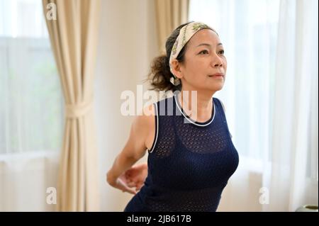 Donna in pensione sportiva asiatica invecchiata che allunga le braccia, facendo yoga, facile allenamento in salotto a casa. Donna sana e anziana. Foto Stock