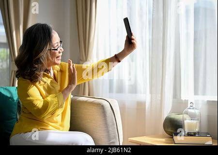 Felice donna asiatica in pensione si siede sul divano, godere di parlare e videochiamare con sua figlia al telefono a casa. Foto Stock