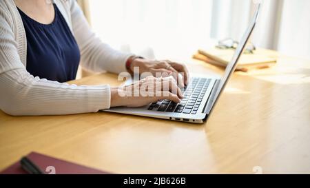 Immagine ritagliata di una donna asiatica di mezza età concentrata seduta alla scrivania e utilizzando un computer portatile. Foto Stock