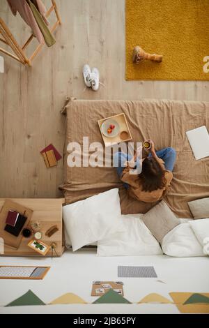 Vista ad angolo alto della signora brunette seduta con gambe incrociate sul letto e bere tè al limone mentre si ha spuntino in camera da letto Foto Stock