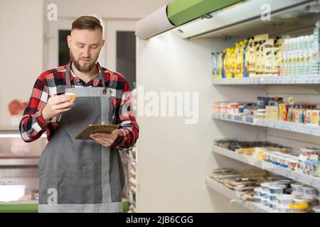 Personale di supermercato maschile che controlla i prodotti utilizzando un tablet. Giovane uomo gioioso, assistente del negozio di alimentari al lavoro, picchiettando sul dispositivo all'interno. Vendita al dettaglio Foto Stock