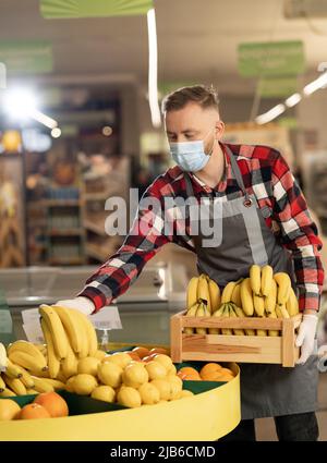 lavoratore del negozio di alimentari maschile che indossa visiera protettiva e grembiule in piedi in negozio e cernita frutta. Bel venditore caucasico in maschera di lavoro in ambienti chiusi Foto Stock