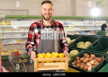 Sorridente lavoratore caucasico in un supermercato con limoni che guardano la macchina fotografica, negozi di alimentari nel reparto frutta e verdura, uomo in grembiule in piedi Foto Stock