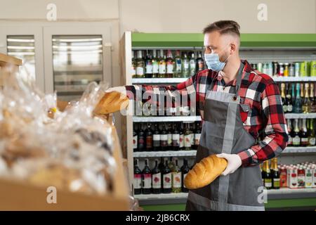 uomo supermercato lavoratore in maschera protettiva mettendo pane fresco sugli scaffali, negozio di alimentari con acqua e dolci, negozio di shopping Foto Stock