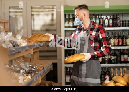 uomo supermercato lavoratore in maschera protettiva mettendo pane fresco sugli scaffali, negozio di alimentari con acqua e dolci, negozio di shopping Foto Stock