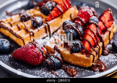 Waffle belgi con cioccolato, fragola, mirtilli e zucchero in polvere su piatto scuro Foto Stock