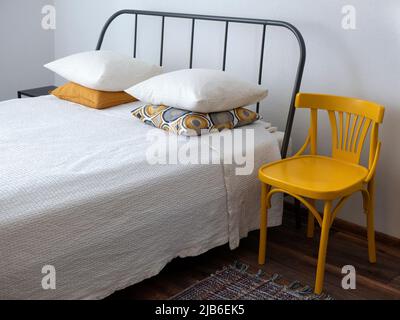 Interno semplice con letto, sedia e tappeto in bianco e giallo. Camera da letto minimalista e luminosa Foto Stock