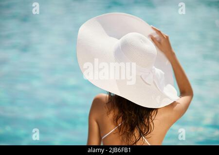 Ragazza graziosa in cappello bianco prendere il sole nella giornata estiva soleggiata. Vista posteriore della signora tenera che indossa un bel cappello con arco che gode la vacanza, con acqua turchese sfocata sullo sfondo. Concetto di vacanza. Foto Stock