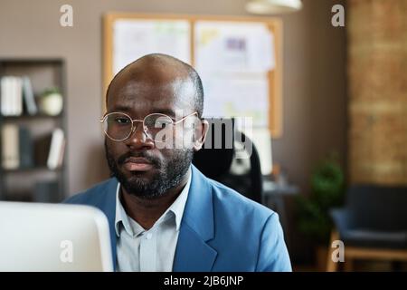 Un uomo d'affari africano serio in occhiali guardando il monitor del computer e concentrandosi sul suo lavoro in ufficio Foto Stock