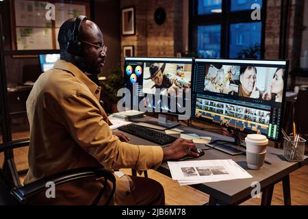 Uomo africano in cuffie seduto sul suo posto di lavoro in ufficio di fronte al monitor del computer e l'editing di foto Foto Stock